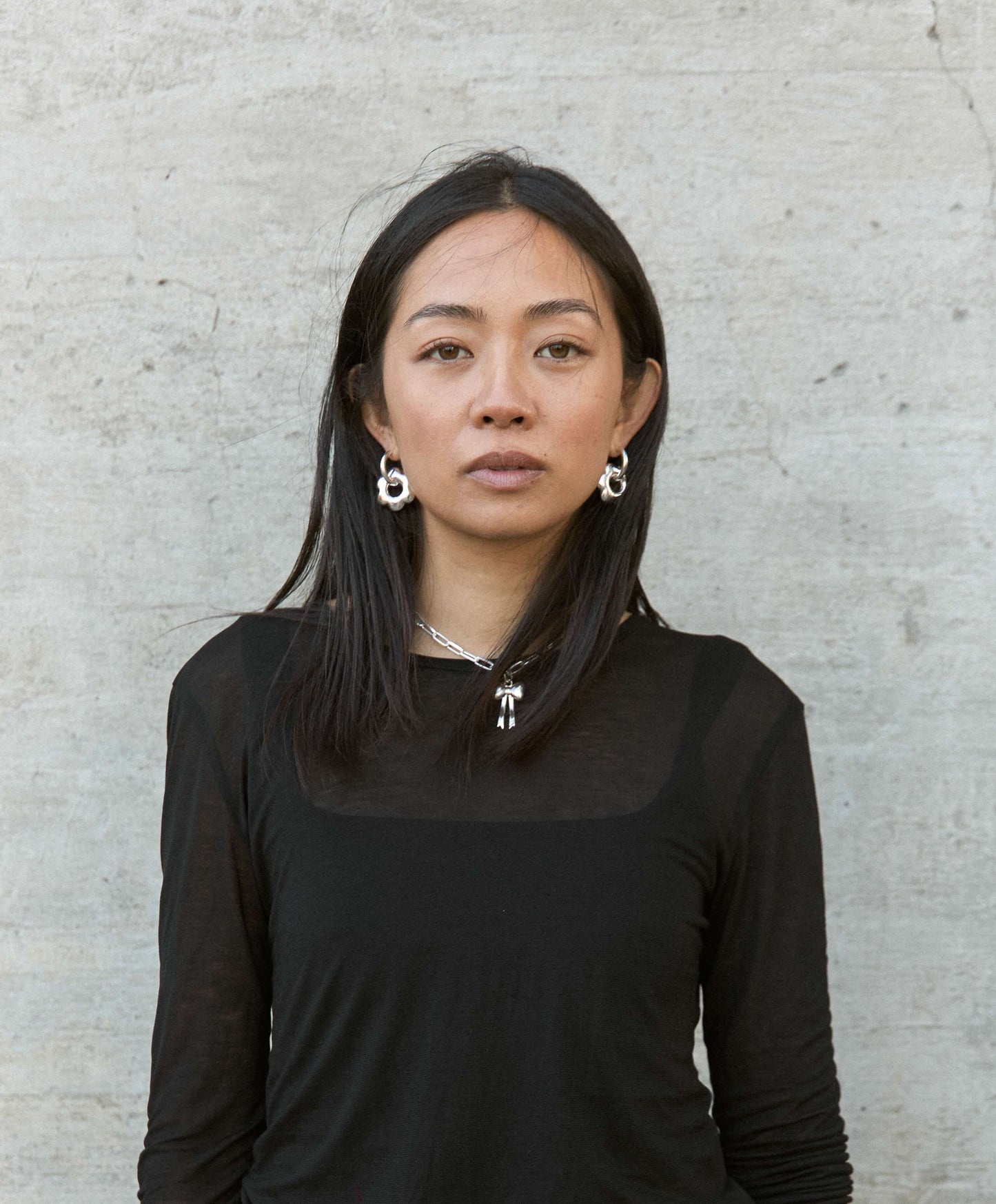 Woman wearing large, puffy flower, silver hoop earrings against a light gray concrete background