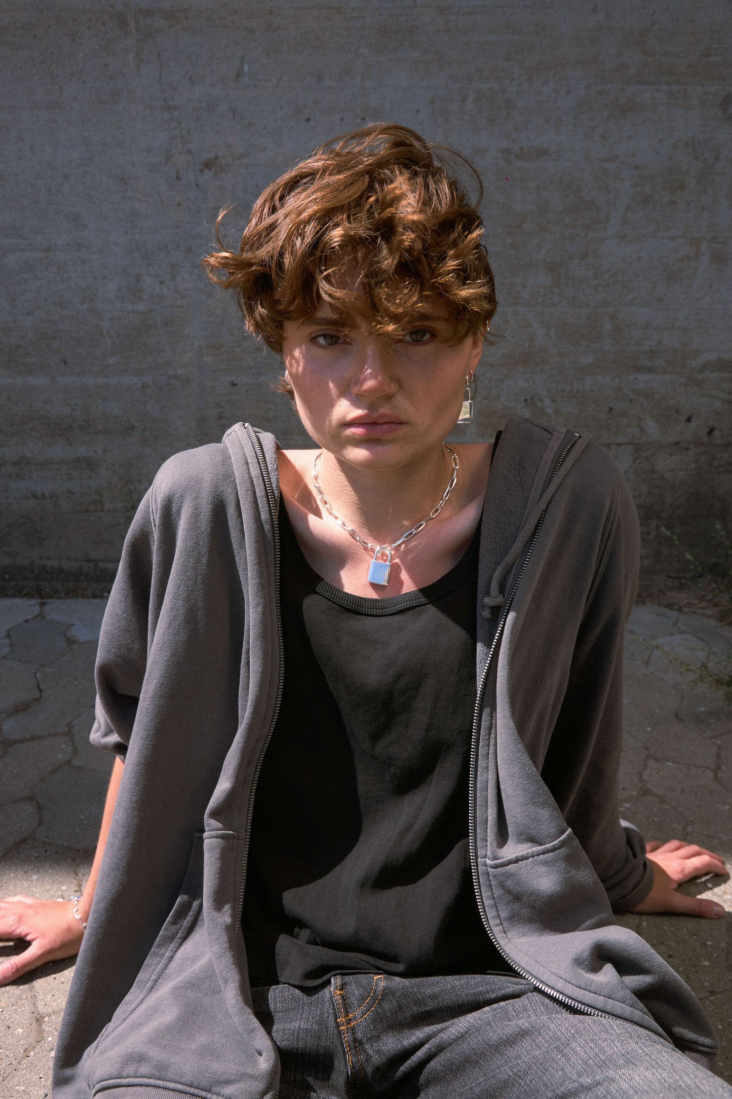 Woman wearing a chunky, silver padlock necklace, gray hoodie and black top sitting against a concrete wall.
