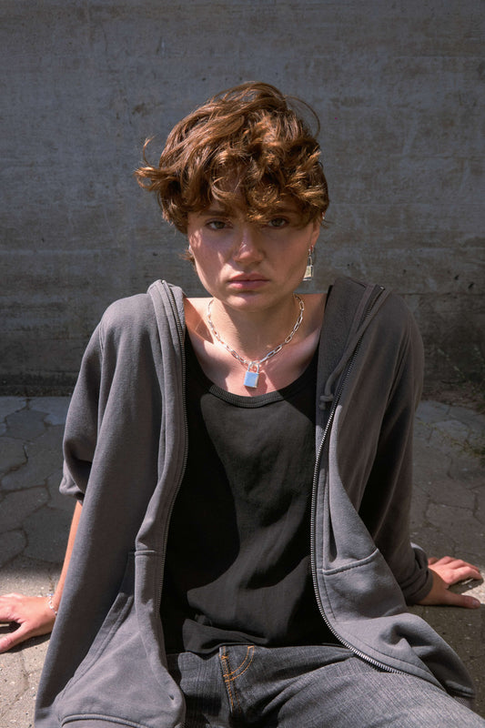 Woman wearing a chunky, silver padlock necklace, gray hoodie and black top sitting against a concrete wall.